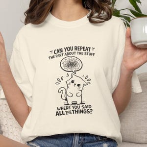 May include: Cream-colored t-shirt with a black graphic of a cat and text. The cat is drawn with a surprised expression, and a thought bubble above its head contains scribbled lines. The text reads "CAN YOU REPEAT THE PART ABOUT THE STUFF" and "WHERE YOU SAID ALL THE THINGS?"