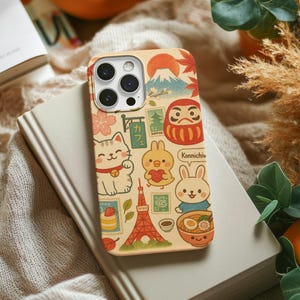 Könnte beinhalten: Eine beige Handyhülle mit japanischen Illustrationen. Die Hülle zeigt den Fuji, eine Daruma-Puppe, eine Glückskatze und andere kulturelle Symbole. Das Design enthält den Text "Konnichiwa" und verschiedene Briefmarken. Die Hülle liegt auf einem Buch.