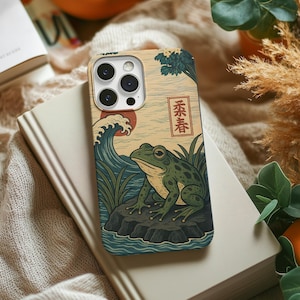 May include: A phone case featuring a Japanese-inspired design with a frog on a rock, waves, and the text "春春". The case has a beige background with a colorful illustration. The phone case is on top of a book.