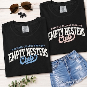 May include: Two black t-shirts with the text "I SURVIVED COLLEGE DROP-OFF EMPTY NESTERS Club" in white and pastel colours. A pair of denim shorts, sunglasses, and earbuds are also in the image. The shirts are labelled "L-G-G".