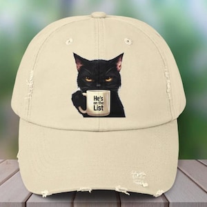 May include: A beige baseball cap with a distressed brim. The cap features a graphic of a black cat holding a mug that reads "He's on the List". The cat has yellow eyes and a stern expression.