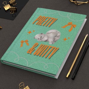 May include: A teal journal with the words "PRETTY & WITTY" in orange lettering, along with a graphic of a white kitten. The journal has a glittery texture and is decorated with orange bows and stars. A gold and black pencil are next to the journal.
