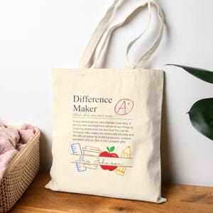 Może przedstawiać: Kremowa torba z napisem "Difference Maker" i definicją wydrukowaną na czarno. Torba posiada grafikę z czerwoną jabłkiem, ołówkiem i linijką. Pod grafiką widnieje napis "Mrs. Johnson".