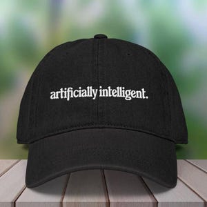Peut inclure: Casquette de baseball noire avec le texte "artificially intelligent" brodé en blanc. La casquette a une visière incurvée et une fermeture à sangle en tissu. Le chapeau est présenté sur une surface en bois.