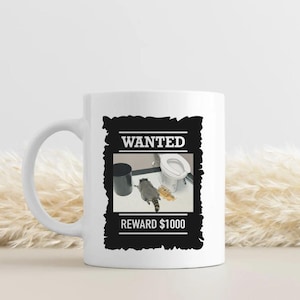 Può includere: Tazza in ceramica bianca con grafica in bianco e nero di un procione in un bagno. Il design include il testo "WANTED" e "REWARD 1000$" in una cornice nera, per un tocco umoristico.