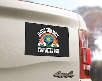 May include: A rectangular black bumper sticker with the text "Have the day you voted for" in white, arched above a rainbow and smiling flowers. The sticker is affixed to the back of a white vehicle.