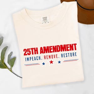 Puede incluir: Camiseta color crema con el texto "25TH AMENDMENT" en rojo y "IMPEACH. REMOVE. RESTORE" en azul. Debajo, tres estrellas y líneas rojas y azules. La camiseta está doblada.