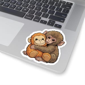 Puede incluir: Una pegatina con dos monos de dibujos animados abrazándose. Un mono es naranja y el otro marrón. La pegatina está en un portátil.