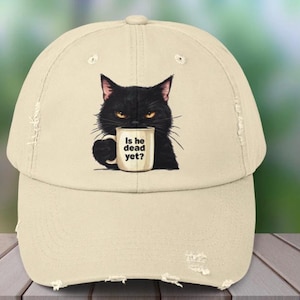 May include: A beige baseball cap with a black cat illustration holding a mug. The mug reads "Is he dead yet?" The cap has a distressed appearance.
