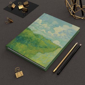 May include: A hardcover journal with a landscape painting on the cover, featuring green hills, blue sky, and white clouds. Gold and black pencils and gold binder clips are arranged around the journal on a dark gray surface.