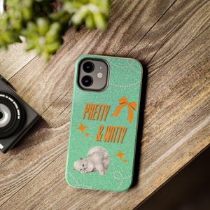 May include: A teal phone case featuring the words "PRETTY & WITTY" in orange, a bow, and a kitten graphic. The case has a glittery finish and is decorated with orange star shapes and a string of pearls.