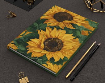 May include: A hardcover journal featuring a vibrant sunflower design. The cover showcases large yellow sunflowers with brown centers and green leaves. Gold and black pencils and gold binder clips are also visible.