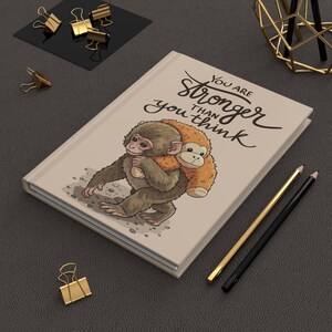 May include: A beige journal with the text "You are stronger than you think" and an illustration of two monkeys. Gold and black pencils and gold binder clips are also visible.