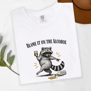 K&ouml;nnte beinhalten: Wei&szlig;es T-Shirt mit dem Text "BLAME IT ON THE ALCOHOL" und einem Cartoon-Waschb&auml;ren mit einem Getr&auml;nk. Der Waschb&auml;r wird mit einer versch&uuml;tteten Tequila-Flasche dargestellt. Das T-Shirt hat einen Rundhalsausschnitt.