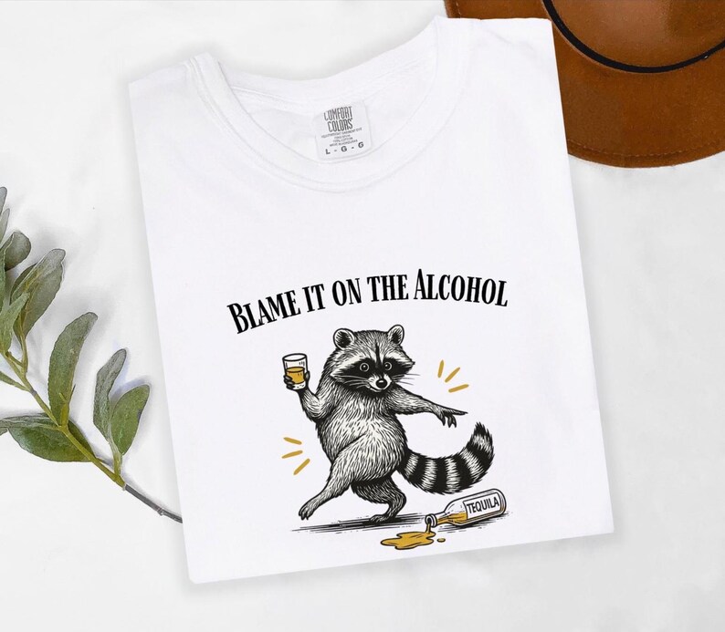 K&ouml;nnte beinhalten: Wei&szlig;es T-Shirt mit dem Text "BLAME IT ON THE ALCOHOL" und einem Cartoon-Waschb&auml;ren mit einem Getr&auml;nk. Der Waschb&auml;r wird mit einer versch&uuml;tteten Tequila-Flasche dargestellt. Das T-Shirt hat einen Rundhalsausschnitt.