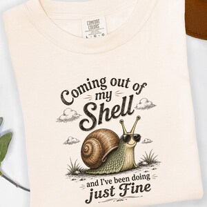 Puede incluir: Camiseta color crema con un gráfico de un caracol con gafas de sol. El texto dice "Coming out of my Shell and I've been doing just Fine." La camiseta está doblada y tiene una etiqueta.