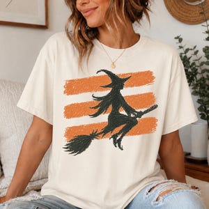 Può includere: Una t-shirt a maniche corte color bianco sporco con una stampa grafica che raffigura la sagoma nera di una strega che vola su una scopa. Il design è sovrapposto a tre strisce orizzontali arancioni, creando un aspetto testurizzato e disegnato.