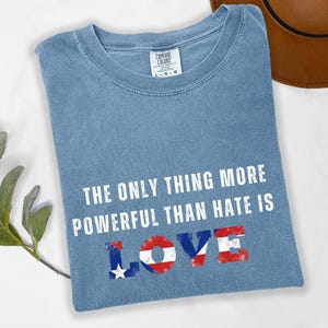 May include: A light blue t-shirt with the text "THE ONLY THING MORE POWERFUL THAN HATE IS LOVE" in white. The word "LOVE" is designed with red, white, and blue accents. The shirt has a Comfort Colors label.
