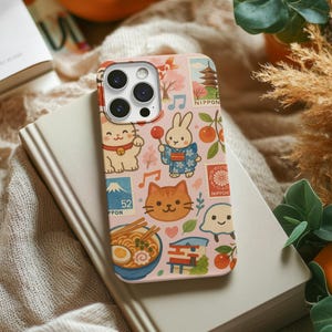 Pode incluir: Uma capa de telefone rosa com um design de tema japonês, com gatos de desenho animado, um coelho em quimono, ramen e selos. A capa tem um recorte para a câmera e a palavra "NIPPON" é visível.