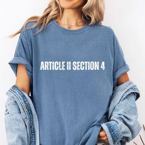 May include: A blue t-shirt with the text "ARTICLE II SECTION 4" in white, distressed lettering. The shirt has short sleeves and a relaxed fit. A denim jacket is draped over the shoulders.
