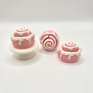 May include: Three pink and white miniature cinnamon rolls. One is on a small white pedestal, another is a single roll, and the third is a double-layered roll. White icing drips down the sides of each roll.