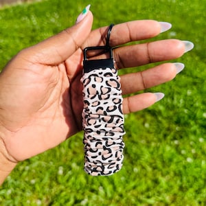 May include: A scrunchie with a black metal clasp and a black key ring. The scrunchie is white with a pattern of black and tan leopard spots. The background is green grass.