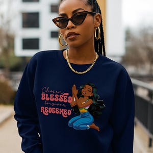 May include: Navy blue sweatshirt with the text "Chosen, Blessed, Forgiven, Redeemed" and an illustration of a woman kneeling in prayer. The woman in the illustration has long green hair, blue jeans, and a colorful top.