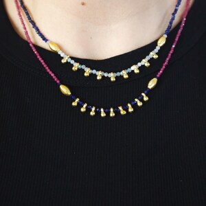 May include: Two layered necklaces with small beads in shades of blue, pink, and gold. The top necklace has light blue beads and gold accents. The bottom necklace has dark blue beads and gold accents. The necklaces are worn against a black background.
