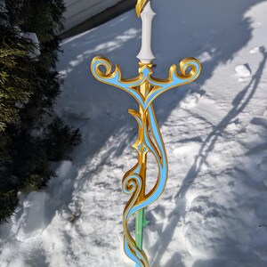 May include: A decorative sword with a gold, light blue, and white design. The sword features a light blue gem at the top, a white handle, and a light green blade. The ornate design includes swirling patterns and geometric shapes.
