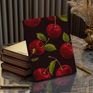 Puede incluir: Una libreta de color rojo oscuro con un patr&oacute;n repetido de cerezas rojas sobre un fondo negro. La libreta est&aacute; cerrada y se encuentra sobre una mesa con un bol&iacute;grafo dorado y una pila de libros.