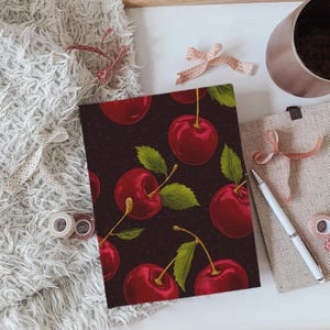 Puede incluir: Una cubierta de cuaderno de color marr&oacute;n oscuro con un patr&oacute;n repetido de cerezas rojas y hojas verdes.
