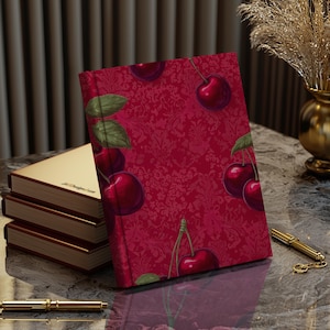 Op de afbeelding: Een rood dagboek met een kersenpatroon. De omslag is dieprood met een herhalend patroon van kersen en bladeren. De kersen zijn donkerrood en de bladeren zijn donkergroen.
