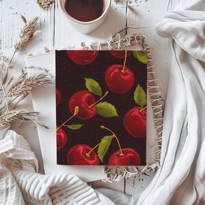 Puede incluir: Una cubierta de cuaderno con un patr&oacute;n repetido de cerezas rojas sobre un fondo negro.