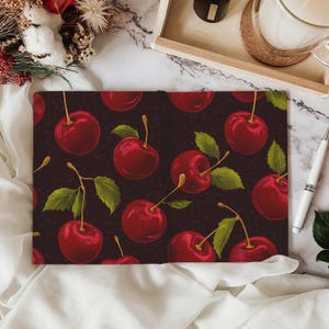 Puede incluir: Una libreta de color rojo oscuro con un patr&oacute;n repetido de cerezas rojas sobre un fondo negro. Las cerezas tienen tallos y hojas verdes.