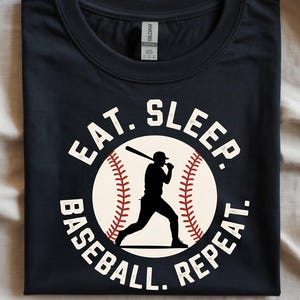 May include: A navy blue t-shirt with a baseball-themed graphic. The design features a baseball with red stitching, a silhouette of a batter, and the words "EAT. SLEEP. BASEBALL. REPEAT." in white lettering.