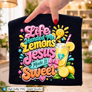 Può includere: Una t-shirt nera con una grafica colorata che riporta il testo Life Handed Me Lemons JESUS Made It SWEET. Il design include un bicchiere di limonata con cannuccia, fette di limone, un fiocco rosa a pois, un cuore con una croce e un sole giallo.