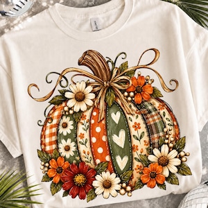 Peut inclure: Un t-shirt couleur crème arborant un grand motif de citrouille décorée de divers motifs, notamment des carreaux, des pois et des cœurs. La citrouille est ornée de fleurs orange, rouges et blanches, de feuilles vertes et d'un nœud décoratif.