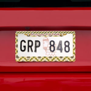 May include: Rectangular license plate with the text "GRP 848" in black, and a decorative border. The plate has a cream background with a design of a building and the text "On my way to la Casita". The plate is mounted on a red surface.