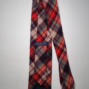 May include: A plaid tie in red, navy, and tan. The tie has a classic pattern and a label that reads "Pendleton". The tie is made of a woven fabric and is suitable for formal or business attire.
