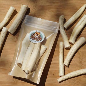 May include: A clear plastic bag filled with several off-white dog treats, some scattered around on a wooden surface. The treats are cylindrical and appear to be dried. A sticker on the bag features a Shiba Inu dog.