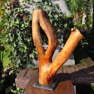 Könnte beinhalten: Handgefertigte Holzskulptur in Form eines Astes, mit einer natürlichen, warmen Farbpalette in Braun- und Orangetönen. Die Skulptur hat eine einzigartige, organische Form und ist auf einem dunkelgrauen Sockel montiert. Ein kleiner Kerzenhalter ist sichtbar.