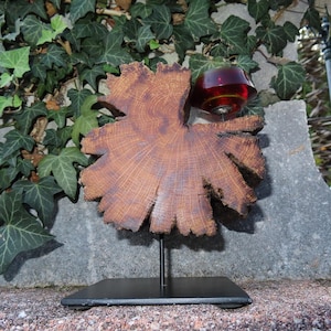 Könnte beinhalten: Eine einzigartige Holzskulptur mit einem großen, braunen, blattförmigen Holzstück, das auf einem schwarzen Metallständer montiert ist. Ein kleines, rotes Glaselement ist am Holz befestigt. Die Skulptur wird im Freien vor dem Hintergrund von grünem Efeu und einer Steinmauer ausgestellt.