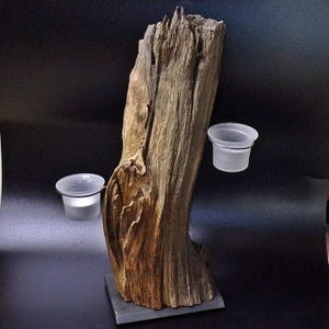 Könnte beinhalten: Ein rustikaler Kerzenhalter aus Holz mit zwei daran befestigten Glaskerzenhaltern. Das Holz ist dunkelbraun und hat ein natürliches, verwittertes Aussehen. Die Kerzenhalter sind aus klarem Glas und mit Metallringen am Holz befestigt.