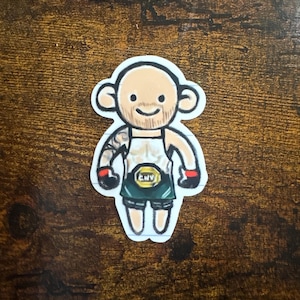 May include: A sticker featuring a cartoon illustration of a bald, smiling figure with a tattoo on the arm. The figure wears a white tank top, green shorts, and a championship belt with the letters "CNV". Red gloves and black wristbands complete the look.