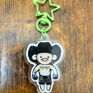 May include: A clear acrylic keychain featuring a cartoon illustration of a fighter wearing a black cowboy hat, black shorts, and blue gloves. The character has a mustache and the text "DREAM BELIEVE MAKE IT HAPPEN" on the chest. It has a lime green star-shaped clasp.