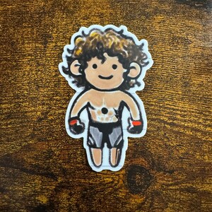 Peut inclure: Autocollant représentant une illustration de dessin animé d'une silhouette musclée avec des cheveux bouclés bruns. La figurine porte des gants noirs et rouges et un short gris. L'autocollant a une bordure blanche.