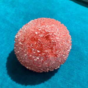 May include: A round, textured, pink decorative object. The object has a crystalline appearance and is set against a bright blue background. The object's surface is uneven, with many small, irregular facets.