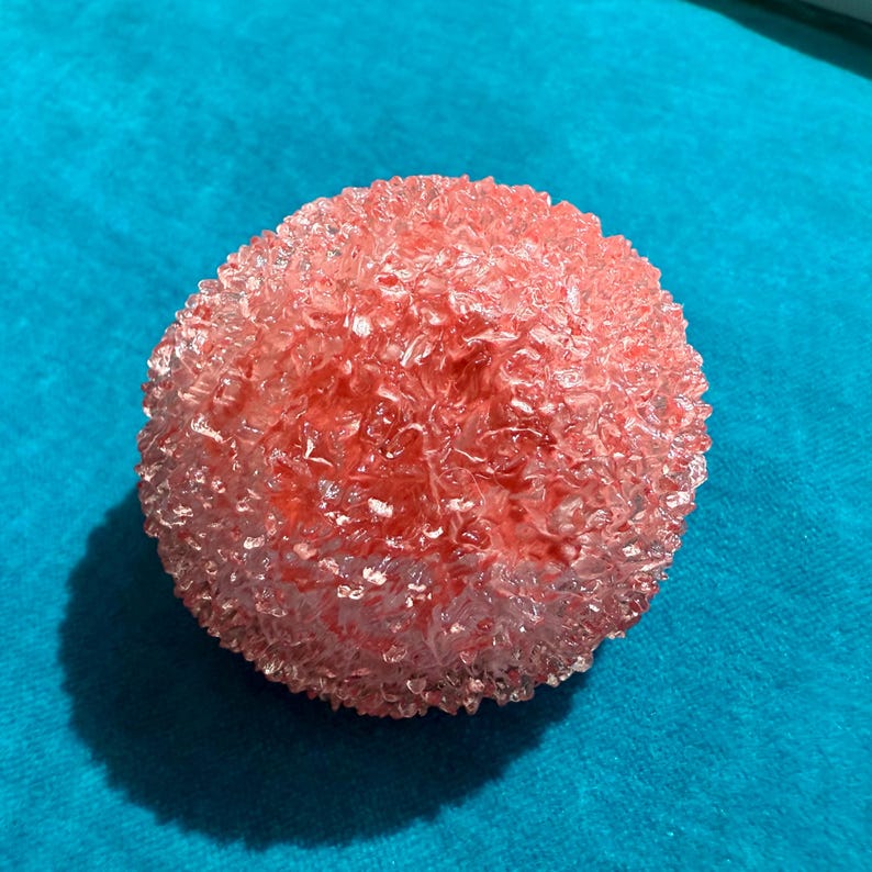 May include: A round, textured, pink decorative object. The object has a crystalline appearance and is set against a bright blue background. The object's surface is uneven, with many small, irregular facets.