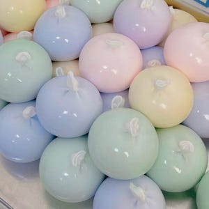 Może przedstawiać: Kolekcja okrągłych przedmiotów w pastelowych kolorach z małym, białym elementem przypominającym sznurek wystającym z góry. Kolory obejmują jasnoniebieski, różowy, zielony, żółty i fioletowy. Przedmioty wydają się gładkie i błyszczące.