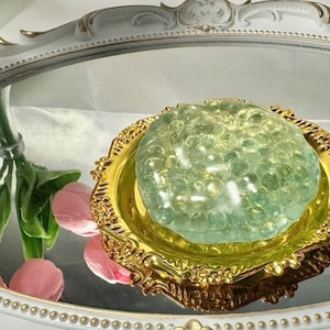 Op de afbeelding: Een groene en gouden slijm met een bubbelstructuur zit op een gouden decoratief dienblad. Het dienblad heeft een sierlijk ontwerp en staat op een wit oppervlak.